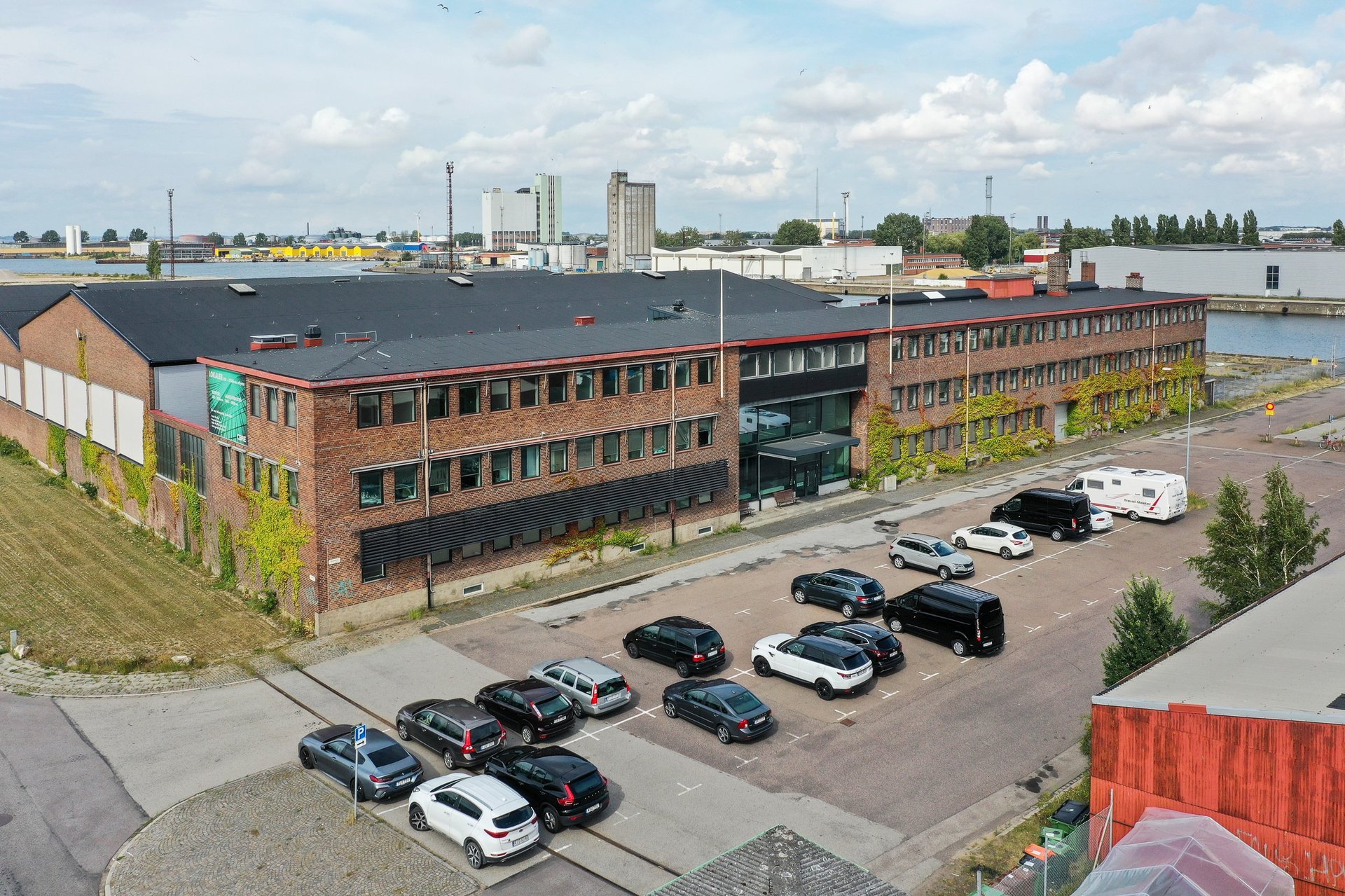 Industriellt hamnområde med tegelmagasin och parkerade bilar i förgrunden, stadssiluett synlig i bakgrunden