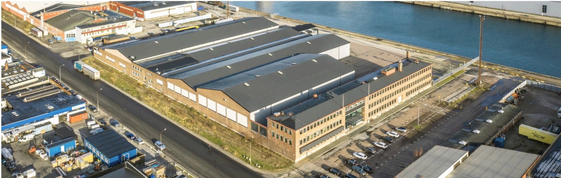 Aerial view of a large industrial waterfront building with solar panels on the roof, docked at a harbor with blue water and nearby structures